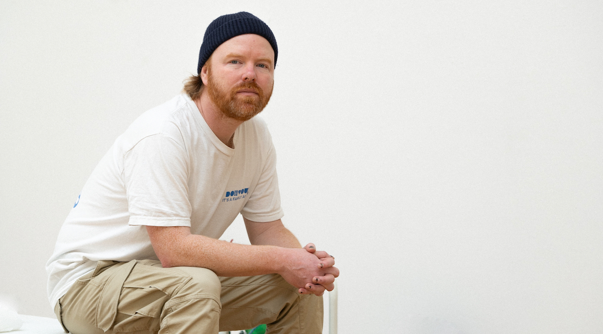 A portrait of Adam Forsythe in his studio. Adam has a ginger beard, a white t-shirt and khaki pants, with a blue beanie on his head, sitting on a bench painting a painting in a green color, against a white background, with studio lighting